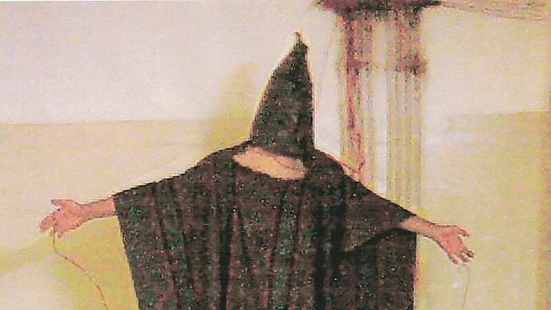 A hooded Iraqi detainee stands in a forced stress position during abuses in Abu Ghraib (Image: WikiMedia Commons)