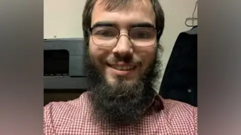 Met Police  Khaled Hussein has brown hair, a large beard and glasses and a red and white shirt - the picture appears to be a selfie and he is smiling into the camera  qhiqquiqdzidqinv