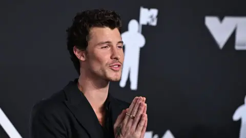 PA Media Shawn Mendes who has dark fairly short wavy hair and stubble. He is wearing a black jacket and looking towards his left with his hands up in prayer qhiddrikeiqzkinv