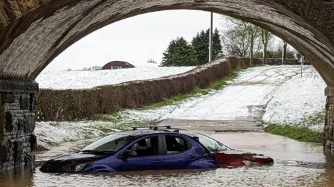 Supplied Two cars - one blue and one red - stuck in flooding under a bridge. Snowy fields and a road can be seen in the background. qhiqhhixdiqzinv