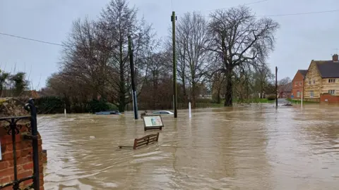 Lesley King Two cars are submerged in floodwater. A bench is also partly under water.