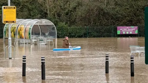 Reece Wilson A woman paddleboards around a flooded supermarket car park. A trolley bay and trollies are partly under water