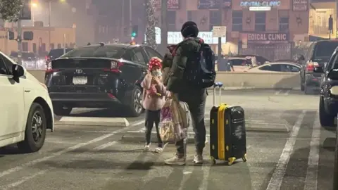 Christal Hayes A woman with her daughter stands on a road with a suitcase in one hand and a bag in another.  qhiqqkiqquierinv