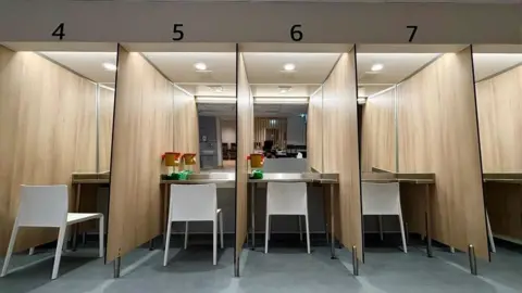 Numbered booths with a white chair in each booth. Each one faces a mirrored wall. It is brightly lit. eiqrriekiqkhinv