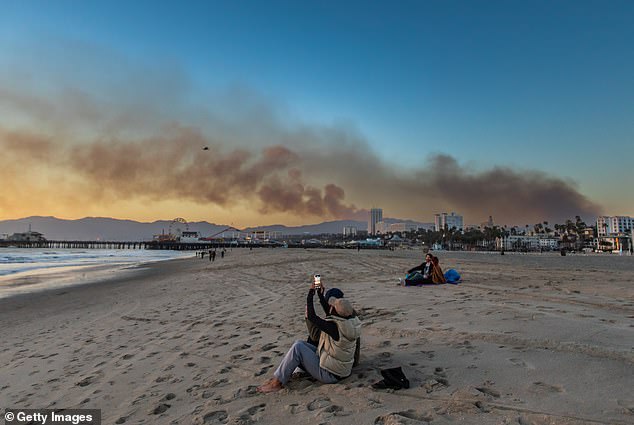 A woman takes a picture on the beach with the Santa Monica pier on the background with smoke from the Palisades Fire