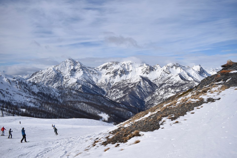 Skiers on a snowy mountain slope overlooking a valley and snow-capped Italian Alps.