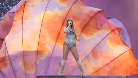 PA Media Taylor Swift on stage at a London concert, in front of a large peach and light purple coloured backdrop. She is singing into a microphone and wearing a one-piece and knee-high boots. eiqeeiqdkiqeqinv