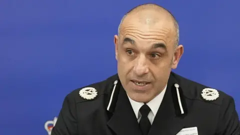 PA Media Thom has short cropped grey and black receding hair and brown eyes. He is wearing a black police uniform blazer with crest logo on both shoulders over a white shirt and black tie. He is talking while looking to the left. eiqrkitritdinv