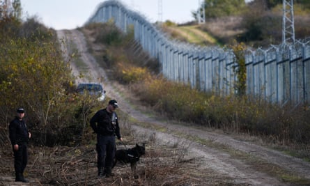 Two policemen in black uniforms, one with a guard dog on a leash, near a metal fence topped by barbed wire in countryside eiqtidqqidtdinv