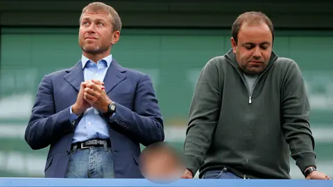 Getty Images Roman Abramovich and Eugene Shvidler standing next to each other. Mr Abramovich is wearing a blue blazer over jeans and has his hands clasped together as he looks into the distance. Mr Shvidler is wearing a quarter-zip jumper in green and is leaning on the railing, looking downwards with a resigned expression dqeiqeriqktidquinv eiqdhidzqiezprw