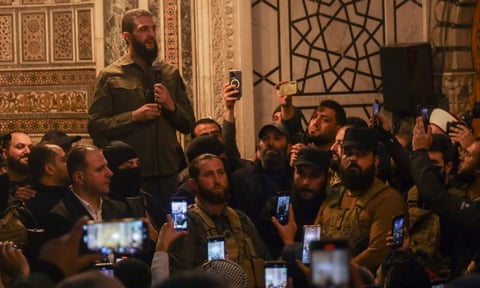 Sharaa speaks to a crowd of men at a mosque: most are wearing khaki shirts and military fatigues, and many in the audience are filming him  qeithiqhdiduinv