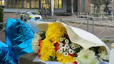 BBC/Simon Thake Flowers and balloons left on the wall outside the school building. A police car and officer can be seen in the background, near the entrance. eiqdhidhiqqkinv