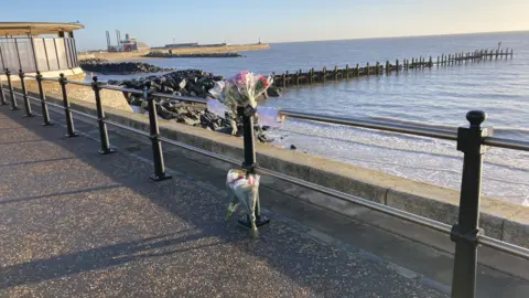 Luke Deal/BBC FLowers and tributes along a sea wall and barrier qhidqhiqtzirzinv