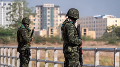 Jonathan Head/BBC Tall buildings in Myanmar’s Myawaddy seen from Thailand’s border, where armed guards stand on a bridge   qhiqhuiduireinv