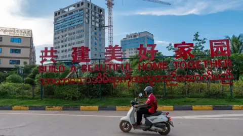 Jonathan Head/ BBC A man riding past a red sign saying ’Let’s build a beautiful home together’  