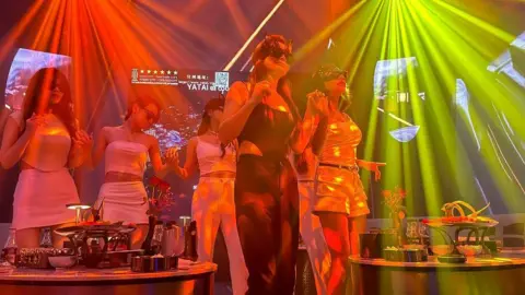 Jonathan Head/BBC Seven women in a karaoke bar, wearing white, black and gold clothing, holding hands and dancing in neon lights