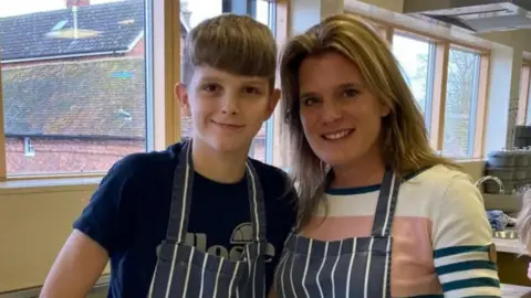 Ellen Roome A woman stands with a young boy. They are both wearing aprons and are smiling. qhidqxiqrdiurinv