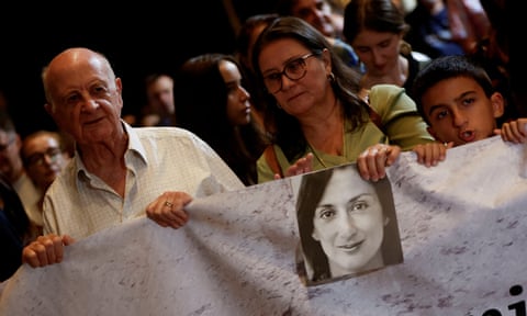 A man, woman and a boy hold a sheet with a b/w picture of Caruana Galizia on it qhiqhhidrkiqeeinv