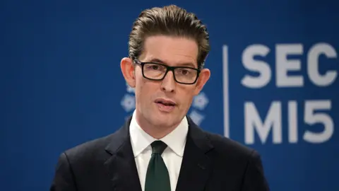 PA Media Ken McCallum, Director General of MI5 - a middle-aged man with slicked back dark brown hair, and wearing dark-rimmed rectangular glasses. He is wearing a dark suit, white shirt and navy blue tie. eiqeriqzridrrinv