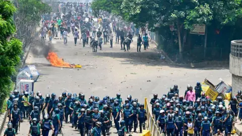 Getty Images A group of police in blue uniforms and helmets, with their backs facing a crowd of protesters. A burning object lies on the ground between them. eiqekiqkdiqztinv