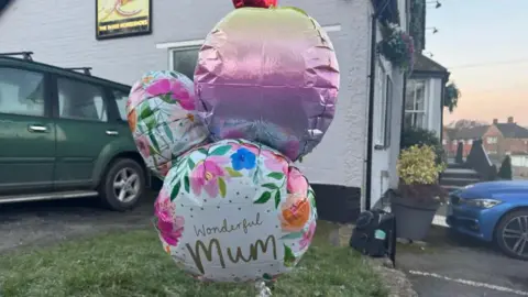 Jo Burn Three balloons on a grass bank in a car park outside a grey pub.