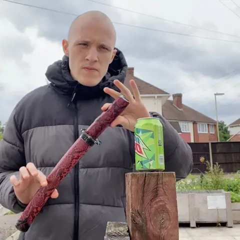 Police handout Petrescu is about to unsheath a sword in a snake-skin holder, to cut a green drinks can on a wooden pole. He has a shaved head and wearing a black winter coat and is standing in what looks like a garden on an estate, with brick houses and planters in the background. 