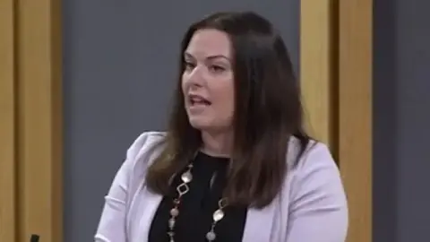 Vikki Howells in the Senedd. She is stood at a lecturn about to speak. She is wearing a black top underneath a lilac jacket and has a beaded and gold necklace on. Her dark shoulder-length hair is untied. qhidquiteiqhinv