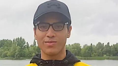 Facebook Nicholas Prosper dressed in glasses, a black baseball hat and yellow and black hoodie while standing in front of a lake. eiqrkidztiezinv