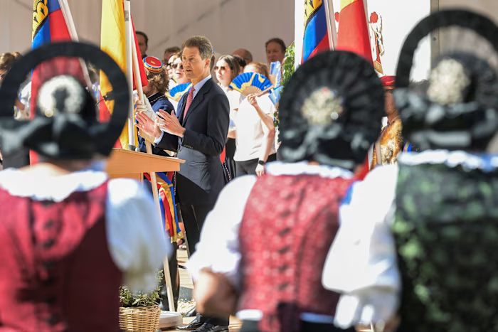 Hereditary Prince Alois speaks at a podium during National Day celebrations, with people in traditional dress and flags in the foreground. qhxidiqxkiqxzinv