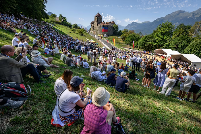 Crowds sit and stand on a grassy hillside below Vaduz Castle during Liechtenstein&rsquo;s National Day celebrations, with tents and flags visible.