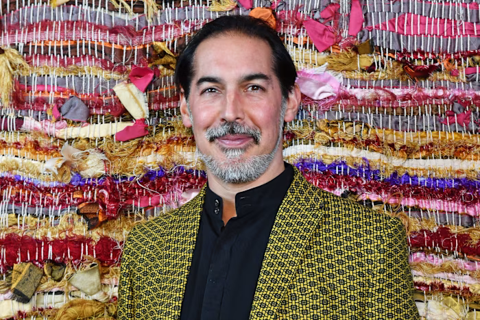Suneil Setiya smiles in front of a colourful woven textile backdrop at a fundraising dinner hosted by Sarabande. qhxidiqxkiqrrinv